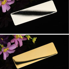 Two rectangular metal nameplates are shown against a black background with purple flowers. The top nameplate is silver, while the bottom one is gold. Both have a smooth, reflective surface perfect for showcasing intricate engraver crafts or precise laser etching designs. These are part of the 20 Packs 4 Colors Stainless Steel Blank Name Tags Badges Nameplate for Laser Engraving by CrealityFalcon.