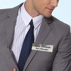 A person dressed in a grey suit, white dress shirt, and blue tie is wearing a name tag on their chest that reads "John Wilson, Sales Manager." The focus is on the upper half of their body, showing only part of their face and torso. The name tag looks professionally crafted, as though made with a precise CrealityFalcon 20 Packs 4 Colors Stainless Steel Blank Name Tags Badges Nameplate for Laser Engraving.