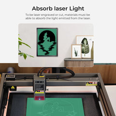 A Creality Falcon laser engraving machine works on a green sheet, carving an image by water. Nearby, a framed print mirrors this design next to plant art and a potted plant. Frosted Colorful Acrylic Sheets (12*12*1/8'') from Creality Falcon are ideal for precise laser cutting and engraving.