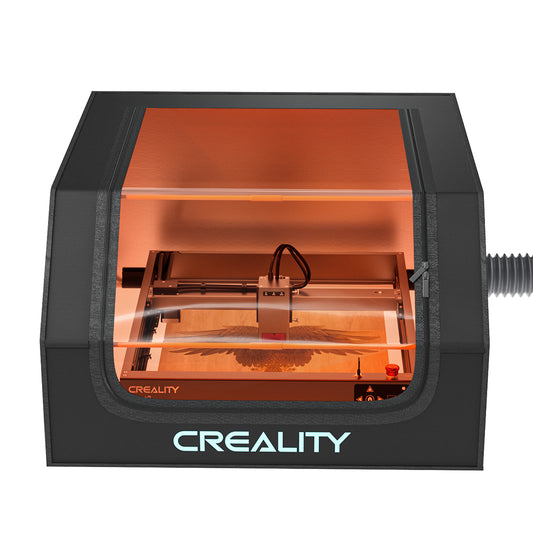 The image showcases a Creality Falcon laser engraver housed within a Falcon Protective Cover for Laser Engraver, featuring an orange viewing window. The machine is actively engraving an intricate design, with light visibly emitted from the laser, while a smoke expel system maintains a clear view and ensures safe operation.