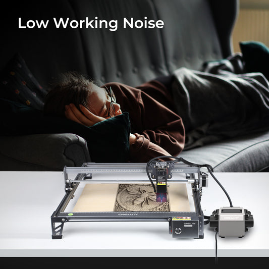 The Creality Falcon's Air Assist for the CR-Laser Falcon 7.5W and 10W Engraver operates quietly on a table, its low working noise maintaining the peaceful atmosphere. In the background, a person relaxes on a couch. The text "Low Working Noise" underscores this feature while ensuring powerful airflow to keep material surfaces clean.