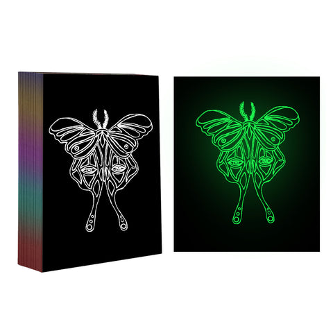 A multi-color stack of books is shown next to a glowing green outline of a moth against a black background. The intricate moth design, crafted with geometric patterns and symmetrical lines, appears as if etched by a CrealityFalcon 50pcs A4 Luminous Scratch Paper Fluorescent Scratch for Falcon Laser Engraving. Two versions are displayed: white on black and green on black.