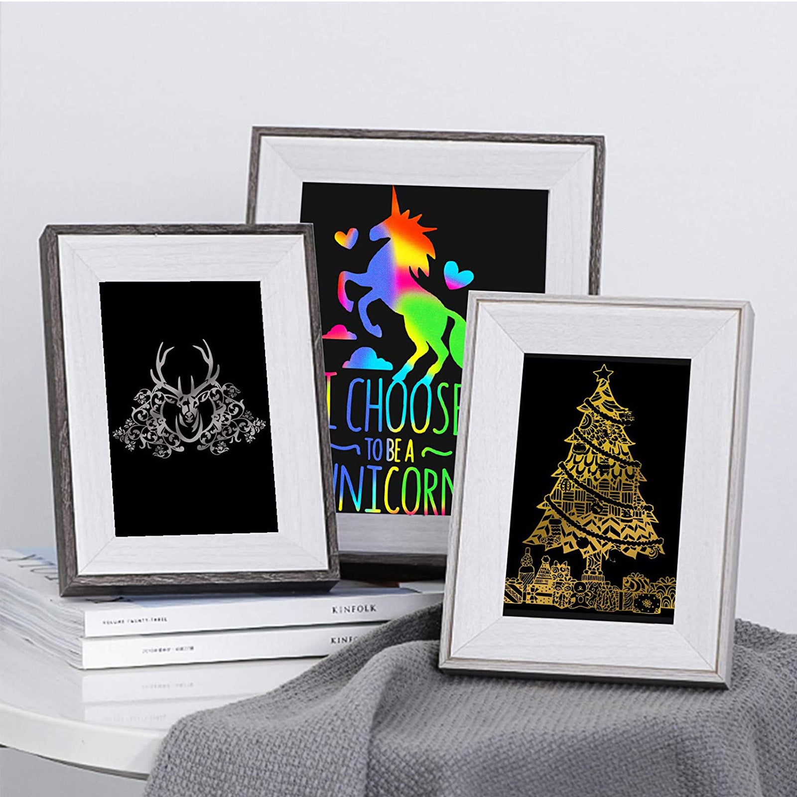 Three decorative framed prints on display. From left to right: an ornate deer head silhouette on black, a vibrant unicorn with the text "I Choose to be a Unicorn," and a festive golden Christmas tree. All frames are set on a light gray surface with a gray cloth below, showcasing exquisite crafts and engraver techniques using 40pcs A4 Gold Silver Scratch Paper Crafts Scratch Arts for Falcon Laser Engraving by CrealityFalcon.