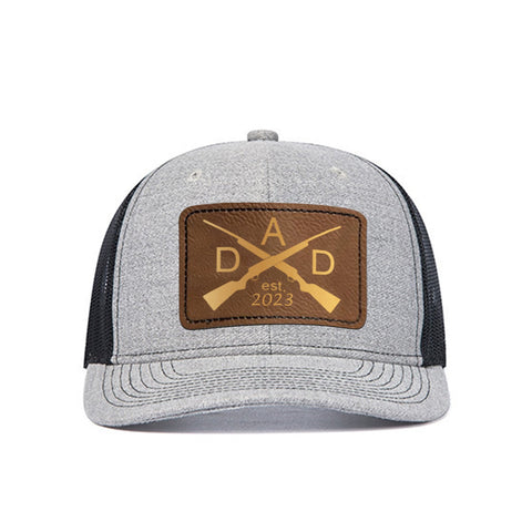 A gray 2 pcs Adjustable Men Mesh Trucker Hat with 4pcs Self-adhesive Cap Stickers for Laser Engraving from CrealityFalcon, featuring a brown leather patch on the front engraved with two crossed rifles and the word "DAD" in large letters, along with "est. 2023" below. The craftsmanship is highlighted by its black mesh sides and back, perfect for any dad who appreciates quality.