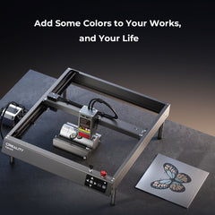 A CrealityFalcon Falcon2 40W Laser Engraver and Cutter is displayed on a dark surface. Next to it lies a metal plate with an engraved, multicolored butterfly design. Above the scene, text reads, "Add Some Colors to Your Works, and Your Life." The advanced safety measures ensure a secure creative process.